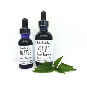 May include: Two glass bottles with dropper tops filled with dark liquid. The bottles are labeled "Rooted Earth Farm Nettle lunar tincture" and are sitting on a white surface with a green nettle leaf.