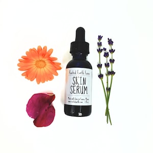 May include: A small, dark glass bottle with a dropper top, labeled "Rooted Earth Farm Skin Serum". The bottle is surrounded by orange and purple flowers on a white background.