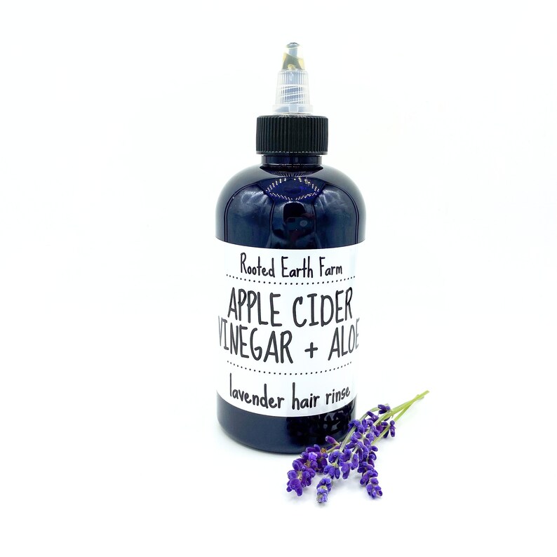 Op de afbeelding: Een donkerblauwe fles met een zwarte dop en een wit etiket met de tekst "Rooted Earth Farm Apple Cider Vinegar + Aloe lavender hair rinse". Op de voorgrond ligt een takje lavendel.