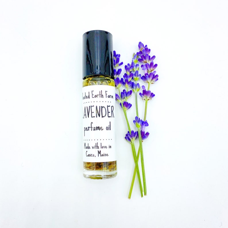 May include: A small glass bottle with a black rollerball top and a white label that reads "United Earth Farm LAVENDER perfume oil Made with love in Casco, Maine". The bottle is sitting on a white surface with a sprig of lavender flowers.