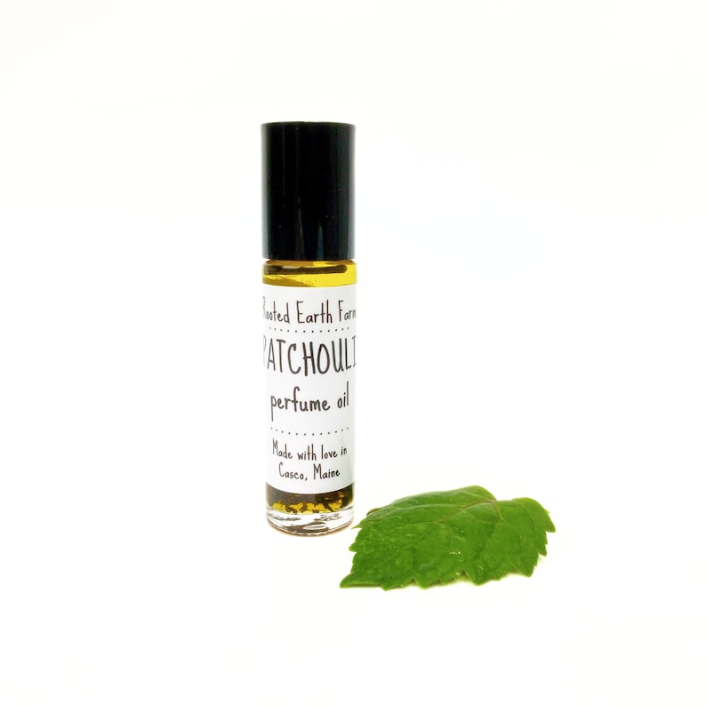 Op de afbeelding: Een klein glazen flesje met patchouli-parfumolie met een zwarte roll-on dop. Het etiket vermeldt "Rooted Earth Farm Patchouli perfume oil" en "Made with love in Casco, Maine".