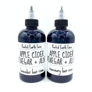 Op de afbeelding: Twee donkerpaarse glazen flessen met zwarte doppen en witte etiketten. De etiketten lezen "Rooted Earth Farm Apple Cider Vinegar + Aloe" met "lavende haar spoeling" en "rozemarijn haar spoeling" onderaan elk etiket.