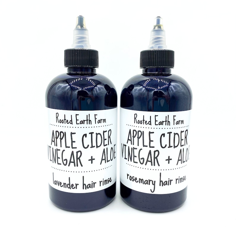 Op de afbeelding: Twee donkerpaarse glazen flessen met zwarte doppen en witte etiketten. De etiketten lezen "Rooted Earth Farm Apple Cider Vinegar + Aloe" met "lavende haar spoeling" en "rozemarijn haar spoeling" onderaan elk etiket.