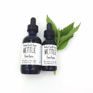 May include: Two glass bottles with dropper tops filled with a dark liquid. The bottles are labeled "Rooted Earth Farm Nettle Tincture" and are made with love in Casco, Maine. The bottles are sitting on a white surface with a green nettle leaf.