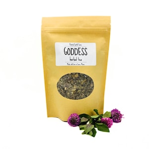 May include: A brown paper bag of "Goddess herbal tea" with a clear window showing the tea blend. The bag is labeled with white text and is accompanied by a sprig of purple flowers.