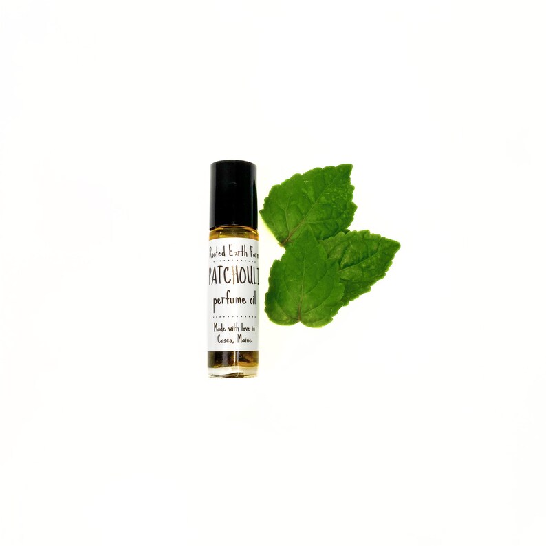Op de afbeelding: Een klein glazen flesje met een zwarte rolbal-applicator en een wit etiket met de tekst "Tinted Earth Farms PATCHOULI perfume oil Made with love in Casco, Maine". Het flesje staat op een witte achtergrond met twee groene bladeren.