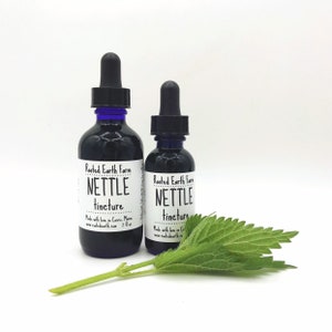 May include: Two glass bottles with blue dropper tops and white labels. The labels read "Rooted Earth Farm Nettle Tincture" and include the website address and size. A green nettle leaf is in the foreground.