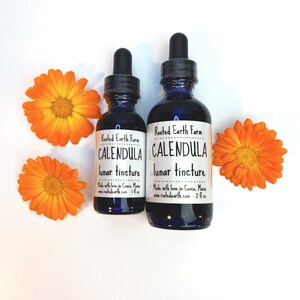 May include: Two dark blue glass bottles with dropper tops, labeled "Rooted Earth Farm Calendula Lunar Tincture". The smaller bottle is 1 fl oz and the larger bottle is 2 fl oz. The bottles are sitting on a white surface with two orange calendula flowers.
