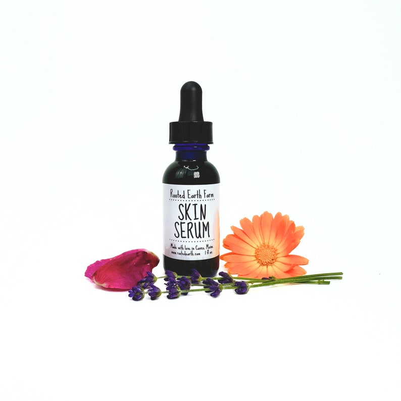 May include: A small, dark brown glass bottle with a blue dropper top. The bottle is labeled "Rooted Earth Farm Skin Serum" and has a white background with a watercolor-like design. The bottle is surrounded by lavender flowers and a single orange flower.