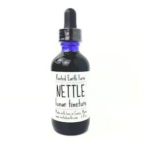 May include: A dark glass bottle with a blue dropper top and a white label that reads "Rooted Earth Farm NETTLE lunar tincture Made with love in Casco, Maine www.rootedearth.com 2 fl oz"