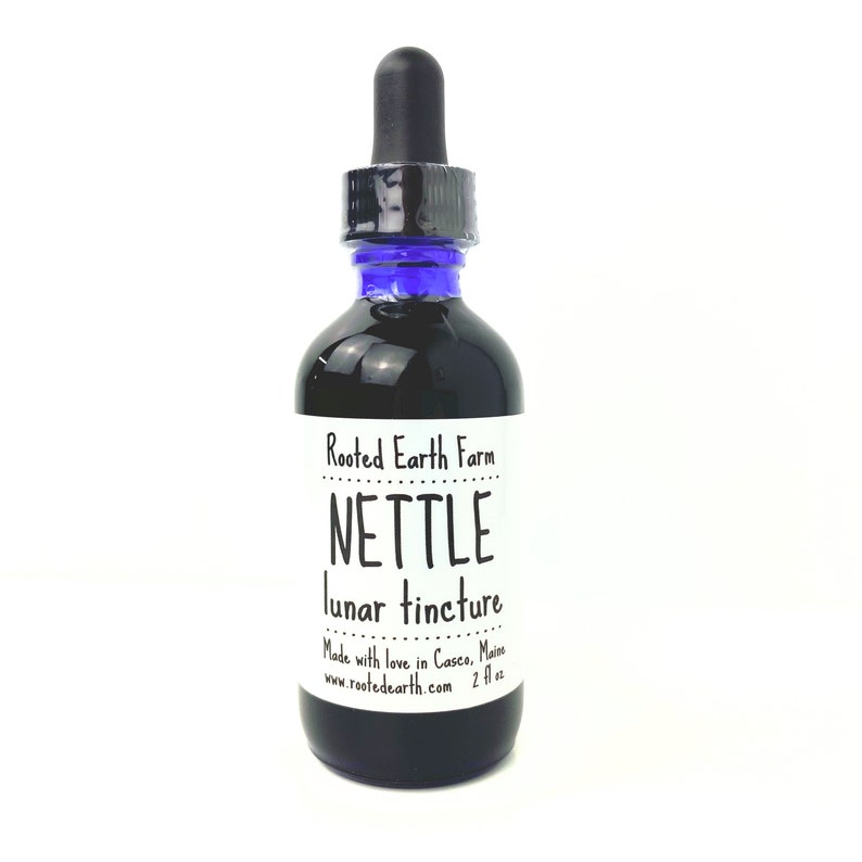 May include: A dark glass bottle with a blue dropper top and a white label that reads "Rooted Earth Farm NETTLE lunar tincture Made with love in Casco, Maine www.rootedearth.com 2 fl oz"