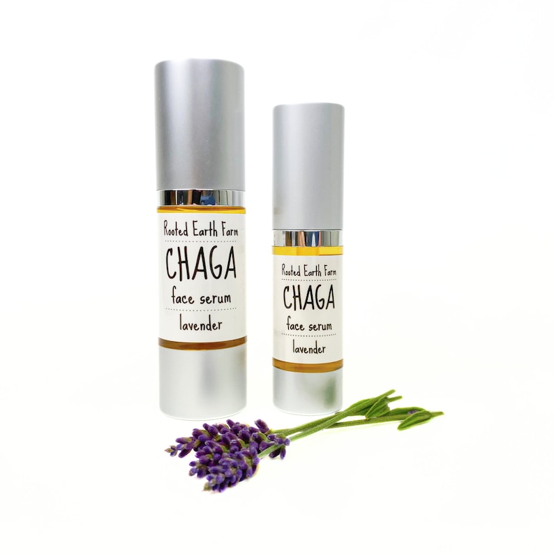Pode incluir: Dois frascos prateados de soro facial Chaga com lavanda da Rooted Earth Farm. Os frascos est&atilde;o sobre uma superf&iacute;cie branca com um ramo de lavanda.