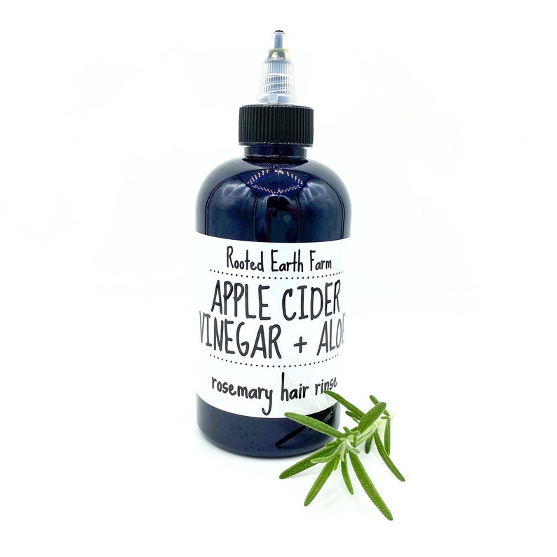 Op de afbeelding: Een donkerblauwe glazen fles met een zwarte dop en een wit etiket met de tekst "Rooted Earth Farm APPLE CIDER VINEGAR + ALOE rosemary hair rinse". Voor de fles ligt een takje rozemarijn.