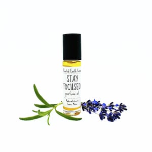 May include: A roll-on perfume oil bottle with a black cap and a white label that reads "Stay Focused". The bottle is surrounded by sprigs of rosemary and lavender flowers. The perfume oil is a light yellow color.