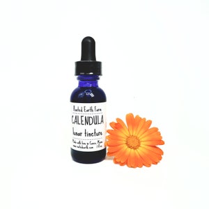 May include: A blue glass bottle with a dropper top, labeled "Rooted Earth Farm Calendula lunar tincture". The bottle is sitting on a white surface next to a bright orange calendula flower.