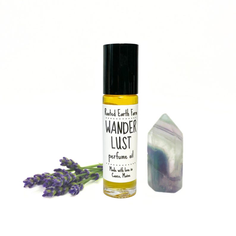 May include: A small glass bottle of perfume oil with a black roll-on top. The label reads "Rooted Earth Farm Wander Lust perfume oil Made with love in Casco, Maine". A purple and green crystal is next to the bottle.