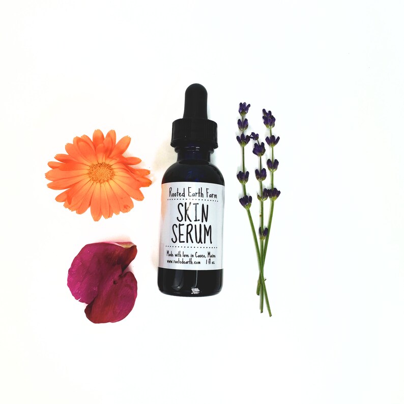 May include: A small dark glass bottle with a dropper top and a white label that reads "Rooted Earth Farm Skin Serum" with the text "Made with love in Cuenca, Maine www.rooted-earth.com 1 fl oz". The bottle is surrounded by three flowers: a purple lavender, a pink rose, and an orange calendula.