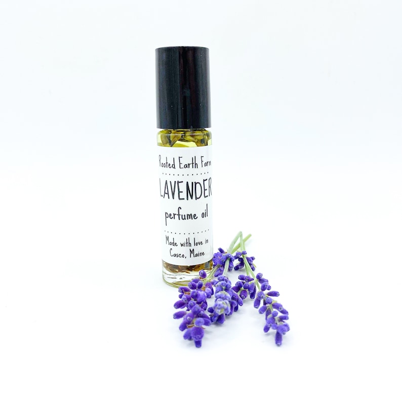 May include: A small glass bottle of lavender perfume oil with a black roll-on top. The bottle has a white label that reads "Rooted Earth Farm LAVENDER perfume oil Made with love in Casco, Maine". The bottle is sitting on a white surface with a few sprigs of lavender flowers.
