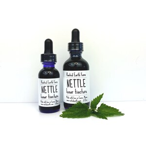 May include: Two glass bottles of nettle lunar tincture, one 1 oz and one 2 oz, with a white label that reads "Rooted Earth Farm" and "Nettle Lunar Tincture". Made with love in Casco, Maine. www.rootedearth.com. A green nettle leaf is in the foreground.