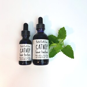 Puede incluir: Dos botellas de vidrio con goteros, ambas etiquetadas como "Rooted Earth Farm Catnip Lunar Tincture". La botella m&aacute;s peque&ntilde;a es de 1 fl oz y la m&aacute;s grande de 2 fl oz. Una ramita de hierba gatera est&aacute; en el fondo.