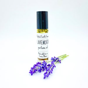 May include: A small glass bottle with a black roll-on top filled with a yellow liquid. The bottle has a white label that reads "Rooted Earth Farm LAVENDER perfume oil Made with love in Casco, Maine". A sprig of purple lavender flowers is in the foreground.