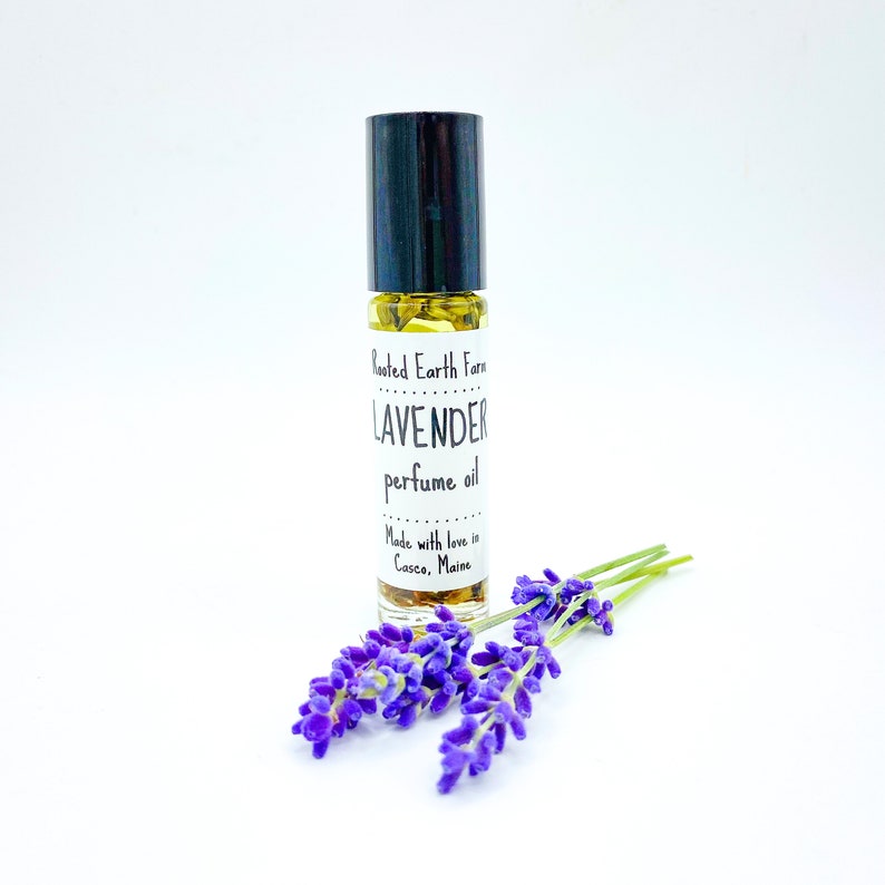 May include: A small glass bottle with a black roll-on top filled with a yellow liquid. The bottle has a white label that reads "Rooted Earth Farm LAVENDER perfume oil Made with love in Casco, Maine". A sprig of purple lavender flowers is in the foreground.