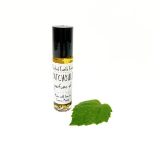 Op de afbeelding: Een klein glazen flesje met een zwarte roll-on dop. Het flesje bevat een gele vloeistof en heeft een wit etiket met de tekst "Rooted Earth Farm Patchouli perfume oil Made with love in Casco, Maine".