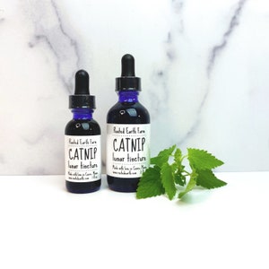 Puede incluir: Dos botellas de vidrio azul con goteros, etiquetadas "Rooted Earth Farm CATNIP lunar tincture". La botella m&aacute;s peque&ntilde;a es de 1 oz y la m&aacute;s grande de 2 oz. Una ramita de hierba gatera verde est&aacute; en primer plano.