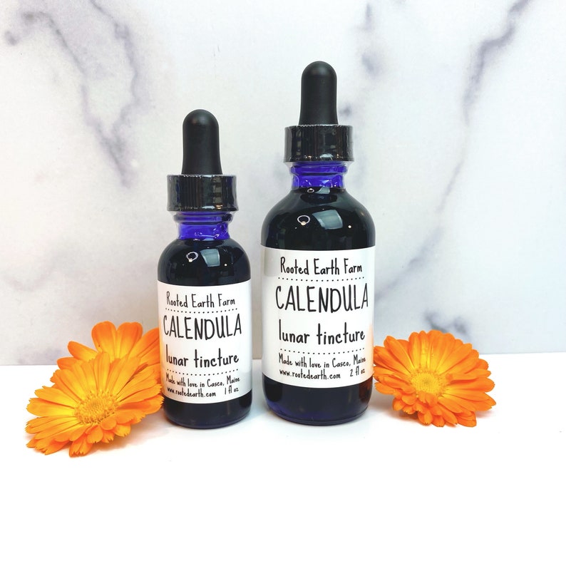 May include: Two glass bottles of Calendula lunar tincture, made by Rooted Earth Farm. The bottles are dark brown glass with blue caps and white labels. The larger bottle is 2 fl oz and the smaller bottle is 1 fl oz.