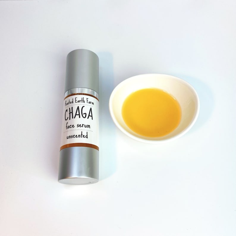Pode incluir: Um frasco prateado com um r&oacute;tulo que diz "Rooted Earth Farm CHAGA face serum unscented". Uma pequena tigela branca com um l&iacute;quido amarelo dentro est&aacute; ao lado do frasco.