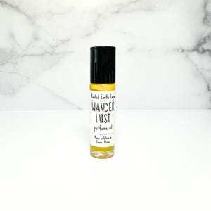 May include: A small glass bottle with a black roll-on top. The bottle contains a yellow liquid and has a white label with the text "Rooted Earth Farm Wander Lust perfume oil Made with love in Gorham, Maine".