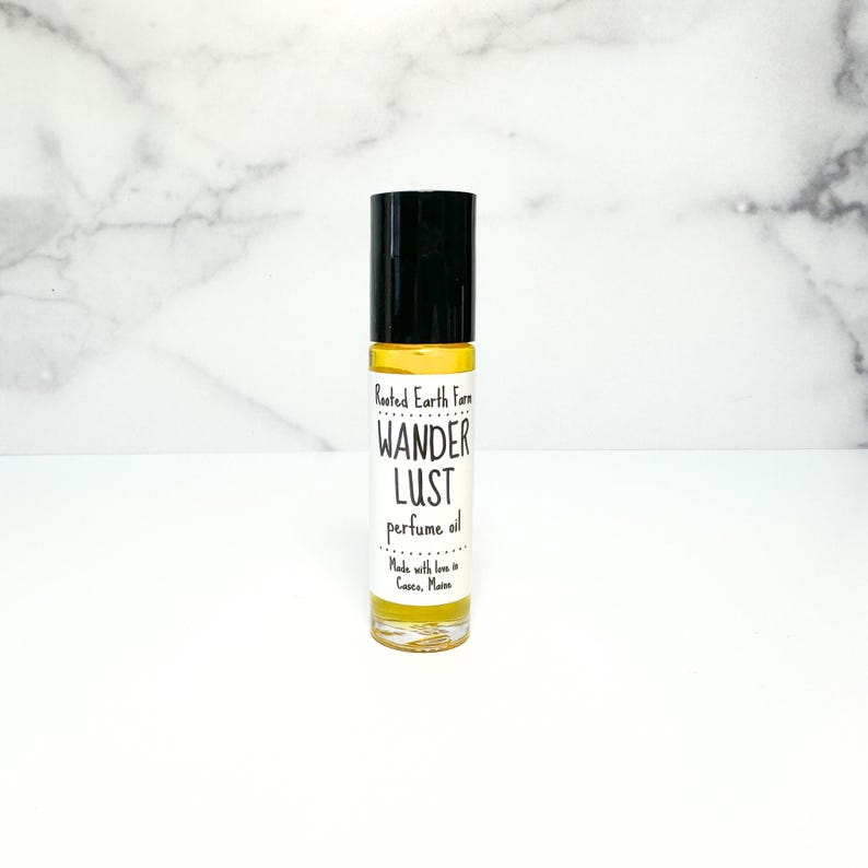 May include: A small glass bottle with a black roll-on top. The bottle contains a yellow liquid and has a white label with the text "Rooted Earth Farm Wander Lust perfume oil Made with love in Gorham, Maine".