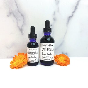 May include: Two bottles of Rooted Earth Farm Calendula lunar tincture. The bottles are dark brown glass with blue caps and white labels. The smaller bottle is 1 oz and the larger bottle is 2 oz. The bottles are sitting on a white marble surface with orange calendula flowers.