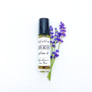 May include: A small glass bottle with a black rollerball top filled with a yellow liquid. The bottle has a white label with the text "United Earth Farm Lavender perfume oil Made with love in Casco, Maine". A sprig of lavender flowers is next to the bottle.