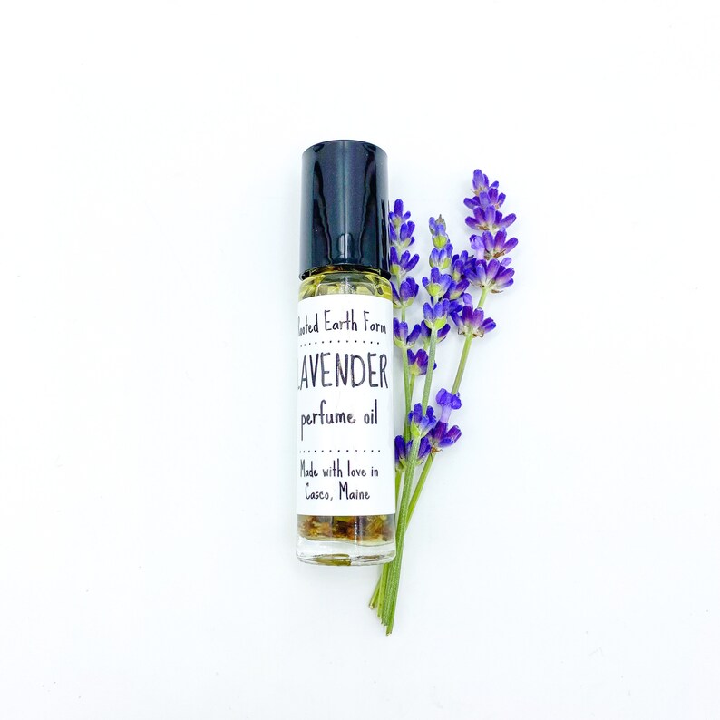 May include: A small glass bottle with a black rollerball top filled with a yellow liquid. The bottle has a white label with the text "United Earth Farm Lavender perfume oil Made with love in Casco, Maine". A sprig of lavender flowers is next to the bottle.