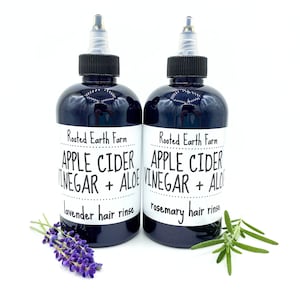 Op de afbeelding: Twee donkerbruine glazen flessen met zwarte doppen en witte etiketten. De etiketten lezen "Rooted Earth Farm Apple Cider Vinegar + Aloe" met "lavende haar spoeling" en "rozemarijn haar spoeling" eronder gedrukt. Een takje lavendel bevindt zich voor de fles aan de linkerkant en een takje rozemarijn voor de fles aan de rechterkant.