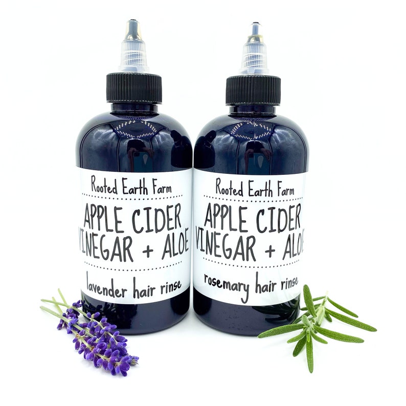 Op de afbeelding: Twee donkerbruine glazen flessen met zwarte doppen en witte etiketten. De etiketten lezen "Rooted Earth Farm Apple Cider Vinegar + Aloe" met "lavende haar spoeling" en "rozemarijn haar spoeling" eronder gedrukt. Een takje lavendel bevindt zich voor de fles aan de linkerkant en een takje rozemarijn voor de fles aan de rechterkant.