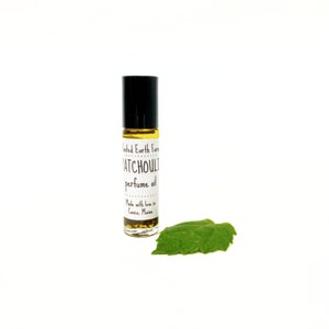 Op de afbeelding: Een klein glazen flesje met een zwarte roll-on-applicator. Het flesje bevat een gele vloeistof en heeft een wit etiket met de tekst "Rooted Earth Farm Patchouli perfume oil Made with love in Casco, Maine". Op de voorgrond ligt een groen blad.