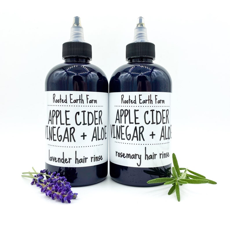 Op de afbeelding: Twee zwarte flessen met witte etiketten. De etiketten lezen "Rooted Earth Farm Apple Cider Vinegar + Aloe" met "lavende haar spoeling" en "rozemarijn haar spoeling" eronder gedrukt. Een takje lavendel bevindt zich naast de fles links en een takje rozemarijn naast de fles rechts.