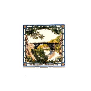 May include: Square ceramic tile featuring a scenic illustration of the Iron Bridge in Shropshire, England, with a river and surrounding greenery. The tile has a decorative border with floral and leaf motifs, and the text "THE IRON BRIDGE SHROPSHIRE 1779".