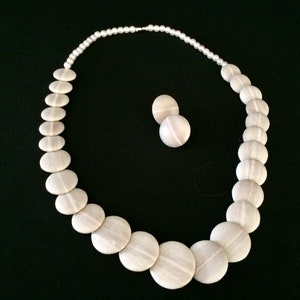 Peut inclure: Un collier composé d'une rangée de petites perles blanches se transformant en disques blancs plats et circulaires avec une texture linéaire subtile, accompagné d'une paire de boucles d'oreilles assorties au motif linéaire blanc.