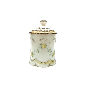 May include: A white porcelain canister with a matching lid, featuring a scalloped rim and base with gold-toned trim. The body is decorated with a delicate floral pattern of yellow roses and green leaves, accented by embossed vertical ridges.