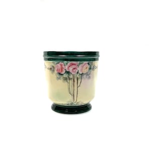 May include: A ceramic vase with a cream-colored body and a dark green rim and base. The vase is decorated with pink roses and green leaves. The roses are painted in a realistic style, and the leaves are detailed. The vase is a decorative item.