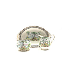 May include: A vintage porcelain tea set featuring a serving platter, a creamer, and a sugar bowl. The set is decorated with a landscape scene of trees and flowers, with gold trim. The pieces are arranged on a white background.