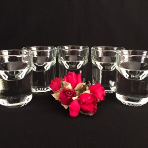 May include: Five clear shot glasses, each containing liquid, are arranged in a line. A small bunch of red roses is positioned in front of the glasses. The background is a plain black.