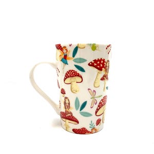 May include: White ceramic mug with a whimsical fairy and mushroom design. The mug features red and white spotted mushrooms, fairies, and teal leaves. The handle is white, and the mug has a tall, cylindrical shape.