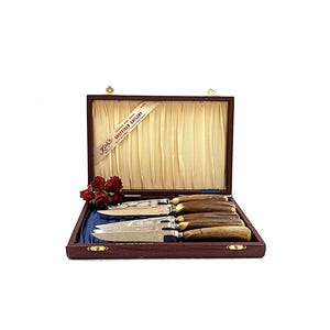 May include: A set of knives in a wooden box. The knives have silver blades and brown handles. The box is lined with a cream-colored fabric and has a ribbon that reads "Sheffield Cutlery". A small bouquet of red flowers is placed near the knives.
