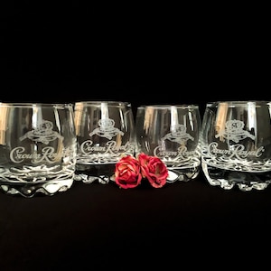 May include: Four clear, lowball glasses with the "Crown Royal" logo etched on the front. The glasses have a rounded shape and a textured base. Two small red flowers are placed in front of the glasses against a black background.