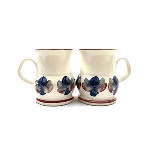 May include: Two cream-coloured ceramic mugs with a rounded base and a handle on the side. Each mug features a hand-painted floral pattern in shades of blue and muted red around the lower section, with a thin red band encircling the base and rim.