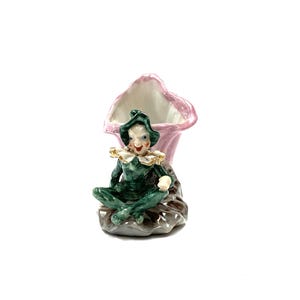 May include: A vintage ceramic figurine of a seated jester in a green outfit and a white ruffled collar. The jester sits in front of a pink and white vase. The base is a dark brown colour. The figurine is on a white background.